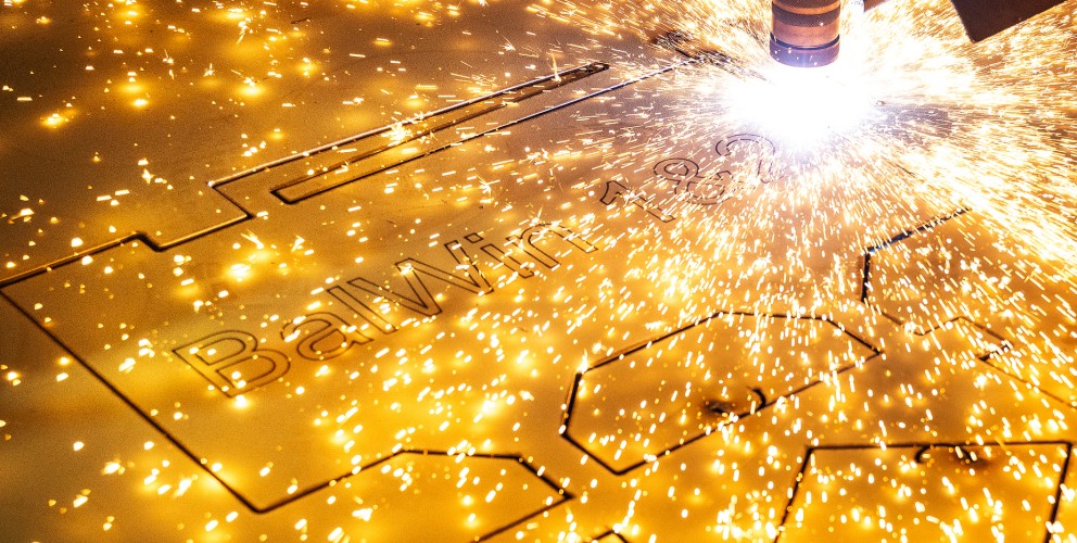 Das Bild zeigt eine Nahaufnahme eines Laserschneiders oder Plasmaschneiders, der in eine goldfarbene Metallplatte schneidet. Ein heller, funkelnder Lichtbogen und zahlreiche leuchtende Funken sprühen von der Schnittstelle. Das intensive Licht dominiert die rechte Seite des Bildes. Die Metallplatte ist horizontal ausgerichtet und weist bereits teilweise ausgeschnittene Formen auf, darunter der Text "BalWin". Der Hintergrund ist unscharf und dunkel, wodurch der Fokus vollständig auf den Schneidevorgang und die Funken gelegt wird. Die Szene vermittelt den Eindruck eines industriellen Fertigungsprozesses mit hoher Präzision und Energie.
English:
The image is a close-up shot of a laser or plasma cutter in action, cutting into a gold-colored metal sheet. A bright, sparkling arc and numerous glowing sparks emanate from the cutting interface. The intense light dominates the right side of the image. The metal plate is oriented horizontally and already shows partially cut-out shapes, including the text "BalWin 3". The background is blurred and dark, putting the entire focus on the cutting process and the sparks. The scene conveys the impression of an industrial manufacturing process characterized by high precision and energy.