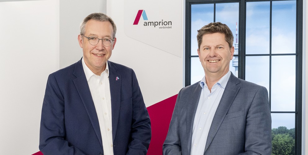 Dr. Christoph Müller und Peter Rüth stehen in Geschäftskleidung in einem Studio. Ihre Hände haben sie jeweils zusammengelegt und locker auf einem weißen Stehtisch abgelegt. Im Hintergrund eine Kulisse mit Amprion Logo und Fenster.
