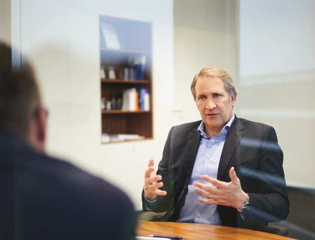 Ein Mann mit grauen Haaren sitzt an einem Konferenztisch in einem modernen Büro. Er trägt ein dunkelblaues Sakko und ein hellblaues Hemd, spricht engagiert und gestikuliert mit beiden Händen. Im Vordergrund sieht man unscharf die Rückenansicht einer weiteren Person. Der Raum ist schlicht eingerichtet, mit einem Regal an der Wand, das einige Bücher und Ordner enthält. Die Atmosphäre wirkt professionell und konzentriert.