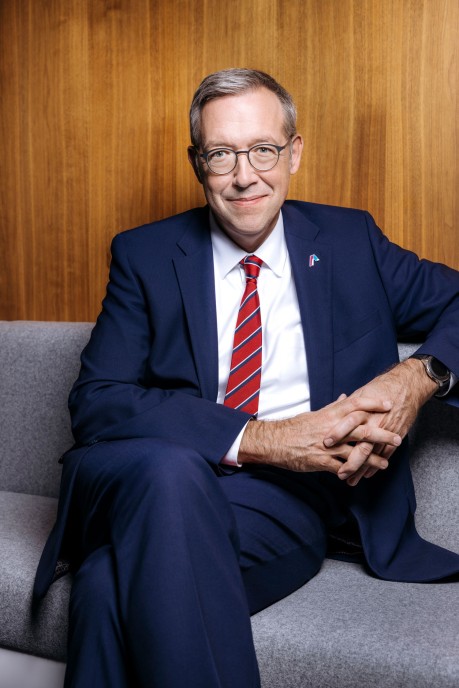 Ein Mann mittleren Alters mit kurzen grauen Haaren und Brille sitzt lächelnd vor einem holzvertäfeltem Hintergrund. Er trägt einen dunkelblauen Anzug, ein weißes Hemd und eine rote Krawatte mit feiner Struktur. Sein Ausdruck wirkt freundlich und professionell.