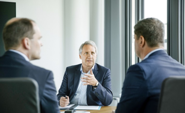 Drei Männer in Anzügen sitzen an einem Konferenztisch in einem modernen, hellen Besprechungsraum. Der Mann in der Mitte, mit grauen Haaren und einem dunkelblauen Sakko, spricht und gestikuliert leicht mit der rechten Hand. Er wirkt konzentriert und führt das Gespräch. Die beiden anderen Männer, deren Gesichter nur von der Seite zu sehen sind, hören aufmerksam zu. Der Raum hat große Fenster, die viel Tageslicht hereinlassen, und der Tisch ist mit Unterlagen, einem Tablet und Stiften bedeckt. Die Atmosphäre wirkt professionell und geschäftlich.