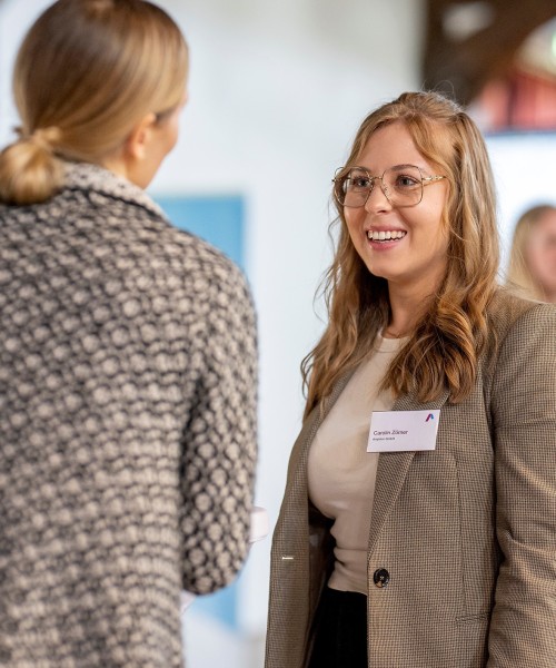 Auf dem Bild sind zwei Frauen zu sehen, die miteinander reden. Die rechte Frau ist von vorne zu sehen und lacht. Sie hat langes, braunes, leicht gewelltes Haar und trägt eine Brille mit einem hellen, filigranen Gestell. Sie hat einen Namensschild an ihrem braun-weiß gemusterten Blazer befestigt. Darunter trägt sie ein helles, einfarbiges Oberteil.
Die zweite Frau steht mit dem Rücken zur Kamera. Sie hat blonde Haare, die zu einem Pferdeschwanz gebunden sind, und trägt eine gemusterte Jacke in dunklen, unscharfen Farben. Der Hintergrund ist hell und unscharf, was den Fokus auf die Frauen lenkt.