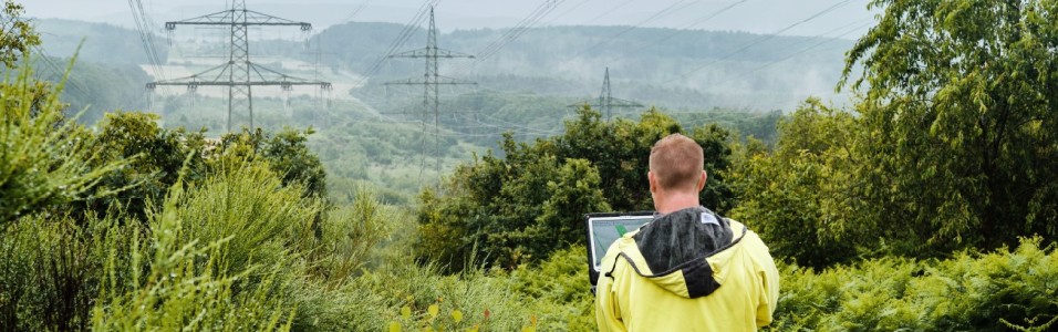 Ein Mann mit gelber Regenjacke steht mit dem Rücken zur Kamera in einer grünen, hügeligen Landschaft und betrachtet Hochspannungsmasten in der Ferne. Er hält ein Tablet oder einen Laptop in den Händen, vermutlich zur Inspektion oder Datenerfassung. Die Masten und Leitungen erstrecken sich durch die Landschaft und verschwinden im Nebel. Dichte Büsche und Bäume rahmen das Bild im Vordergrund ein, während das Wetter feucht und neblig erscheint.