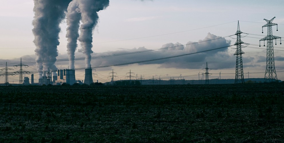 Dieses eher dunkel gehaltene Foto im Querformat zeigt den Blick über einen abgeernteten Acker auf ein Kraftwerk. Aus den Kühltürmen steigt Dampf auf. Im Vordergrund des Kraftwerks eine Stromleitung. 