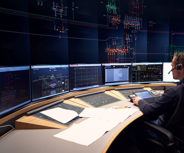 Das Bild zeigt einen Mann mit Headset, der in einem Kontrollraum an einem großen, gebogenen Schreibtisch sitzt. Vor ihm befinden sich mehrere Bildschirme, die technische Diagramme und Daten anzeigen, vermutlich zur Überwachung von Energie- oder Stromnetzen. Im Hintergrund erstreckt sich eine große digitale Anzeige an der Wand, die komplexe Netzstrukturen und Leitungswege in farbigen Linien darstellt. Auf dem Schreibtisch liegen Notizen und Unterlagen