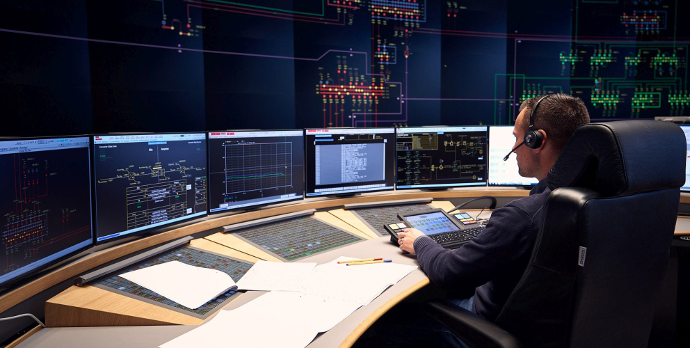 Das Bild zeigt einen Mann mit Headset, der in einem Kontrollraum an einem großen, gebogenen Schreibtisch sitzt. Vor ihm befinden sich mehrere Bildschirme, die technische Diagramme und Daten anzeigen, vermutlich zur Überwachung von Energie- oder Stromnetzen. Im Hintergrund erstreckt sich eine große digitale Anzeige an der Wand, die komplexe Netzstrukturen und Leitungswege in farbigen Linien darstellt. Auf dem Schreibtisch liegen Notizen und Unterlagen