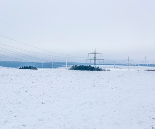 Das Bild zeigt eine verschneite Landschaft mit einer großen, weißen Schneedecke im Vordergrund. Im Hintergrund sind Strommasten mit zahlreichen Leitungen zu sehen, die sich quer durch die Landschaft ziehen. Weiter hinten stehen Windräder, die sich sanft in die Landschaft einfügen. Die Stimmung ist ruhig und winterlich, der Himmel ist grau und bewölkt, wodurch die gesamte Szene kühl und gedämpft wirkt. 