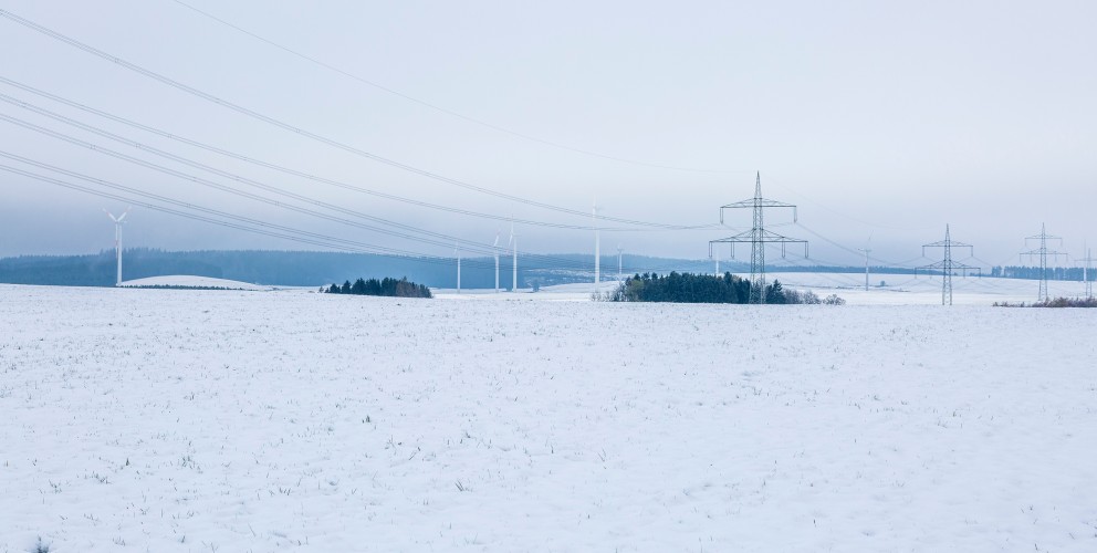 Das Bild zeigt eine verschneite Landschaft mit einer großen, weißen Schneedecke im Vordergrund. Im Hintergrund sind Strommasten mit zahlreichen Leitungen zu sehen, die sich quer durch die Landschaft ziehen. Weiter hinten stehen Windräder, die sich sanft in die Landschaft einfügen. Die Stimmung ist ruhig und winterlich, der Himmel ist grau und bewölkt, wodurch die gesamte Szene kühl und gedämpft wirkt. 