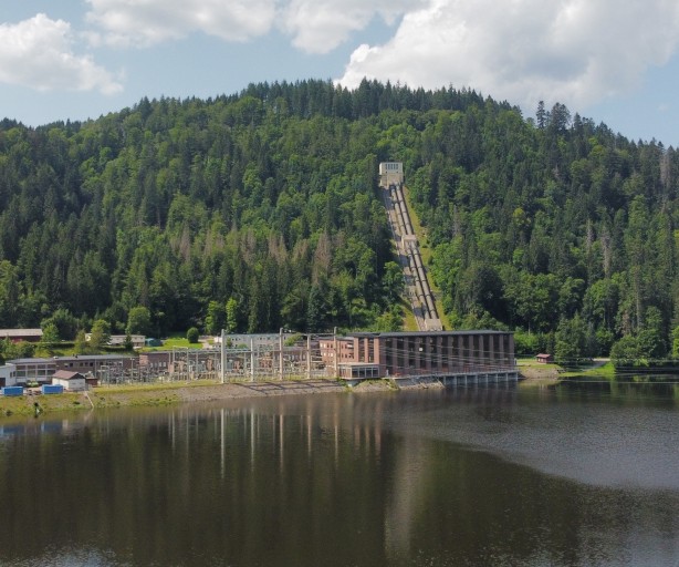 Das Bild zeigt einen See, der von einem dichten Wald umgeben ist. Im Hintergrund sind bewaldete Hügel zu sehen. Am Ufer des Sees steht ein großes Gebäude, das wahrscheinlich ein Kraftwerk ist. Von diesem Gebäude führt ein langer, schmaler Steg aus Metall in den See. Der Himmel ist blau und es sind einige Wolken zu sehen. Die Sonne scheint und es ist ein schöner Sommertag. Das Wasser des Sees ist still und spiegelt die umliegende Landschaft wider.