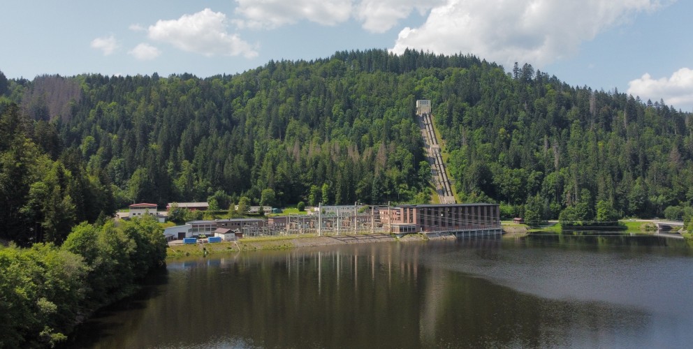 Das Bild zeigt einen See, der von einem dichten Wald umgeben ist. Im Hintergrund sind bewaldete Hügel zu sehen. Am Ufer des Sees steht ein großes Gebäude, das wahrscheinlich ein Kraftwerk ist. Von diesem Gebäude führt ein langer, schmaler Steg aus Metall in den See. Der Himmel ist blau und es sind einige Wolken zu sehen. Die Sonne scheint und es ist ein schöner Sommertag. Das Wasser des Sees ist still und spiegelt die umliegende Landschaft wider.