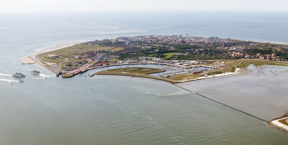 Das Bild zeigt eine luftbildaufnahme der Insel Norderney. Im Vordergrund ist ein Teil des Hafens zu sehen, mit mehreren Anlegestellen für Schiffe. Die Insel selbst ist von Wasser umgeben und erstreckt sich in die Ferne.