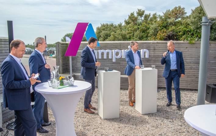 Das Bild zeigt eine Gruppe von Männern in Anzügen, die sich bei einer Outdoor-Veranstaltung oder Präsentation von Ampion unterhalten. Im Hintergrund ist ein großes Ampion-Logo zu sehen, das auf einen Technologie- oder Innovationskontext hindeutet. 