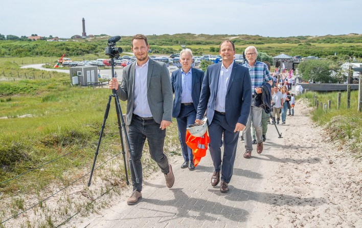 Das Bild zeigt eine Gruppe von Männern, die in einer Küstenlandschaft entlanggehen. Sie tragen Anzüge und scheinen in ein Gespräch vertieft zu sein. Einer von ihnen hält eine Kamera oder ein Videokameraequipment in der Hand.