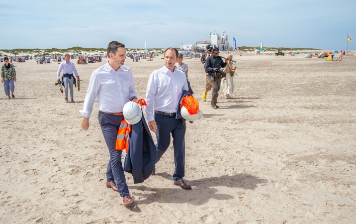 Das Bild zeigt zwei Männer in Bürokleidung, die auf einem Strand spazieren gehen. Sie tragen Schutzhelme und Orange-Warnwesten.
Im Hintergrund ist ein belebtes Strandgelände mit Strandkörben, Fahnen und Gebäuden zu sehen. 