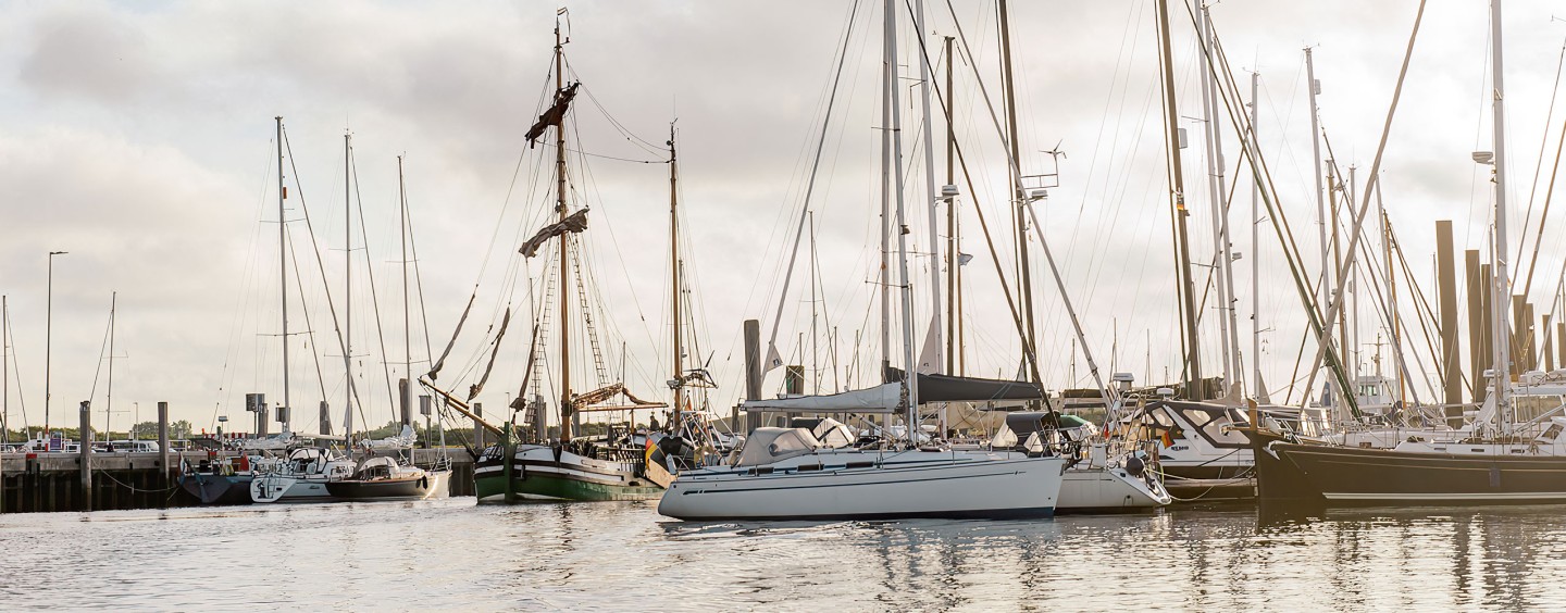 Das Bild zeigt einen Hafen oder eine Marina, die mit verschiedenen Segelbooten und Yachten gefüllt ist. Die Boote sind an den Stegen vertäut, wobei ihre Masten und Takelage deutlich sichtbar sind. Die Szene hat eine bedeckte, etwas düstere Atmosphäre, wobei der bewölkte Himmel sich auf der ruhigen Wasseroberfläche reflektiert. Auch wenn das Wetter nicht ideal zu sein scheint, vermittelt das Bild dennoch ein Gefühl von maritimer Aktivität und der friedlichen Atmosphäre eines Hafenumfelds.