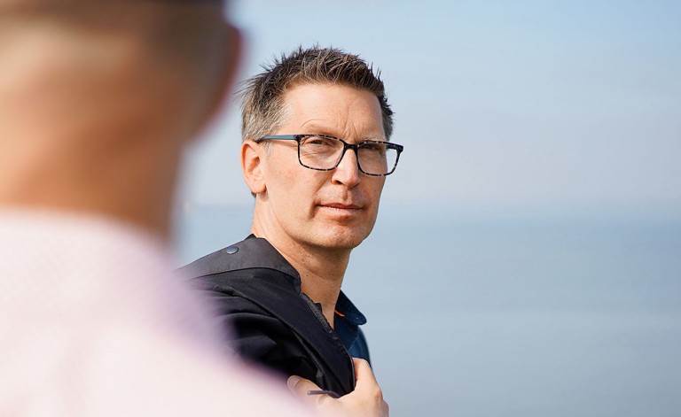 Ein Mann mit kurzen, dunkelblonden Haaren und einer schwarzen Brille schaut ernst in die Kamera. Er trägt ein schwarzes Jackett über der Schulter und steht vor einem ruhigen, verschwommenen Hintergrund aus Himmel und Wasser. Im unscharfen Vordergrund ist die Schulter eines anderen Mannes zu sehen, der ein helles Hemd trägt.
