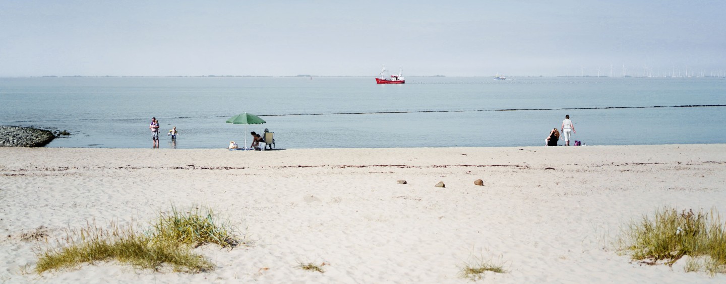 Ein ruhiger Sandstrand mit mehreren Menschen, die die Küste genießen. Ein grüner Sonnenschirm und ein paar Stühle stehen in der Nähe des Wassers. Links stehen Personen im flachen Wasser, während rechts zwei Menschen auf dem Sand sitzen. Im Hintergrund fährt ein rotes Schiff über das ruhige Meer. Der Horizont ist leicht neblig, und am rechten Rand sind Windräder zu erkennen. Der Himmel ist klar und hell.