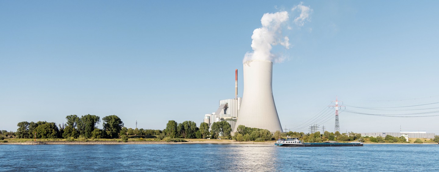 Ein großes Kraftwerk mit einem hohen Kühlturm, aus dem dichter weißer Dampf aufsteigt, steht am Ufer eines Flusses. Vor dem Kraftwerk befinden sich Bäume und eine schmale Landzunge. Ein Frachtschiff fährt auf dem Fluss entlang. Strommasten und Hochspannungsleitungen verlaufen rechts im Bild, während der Himmel klar und blau ist.