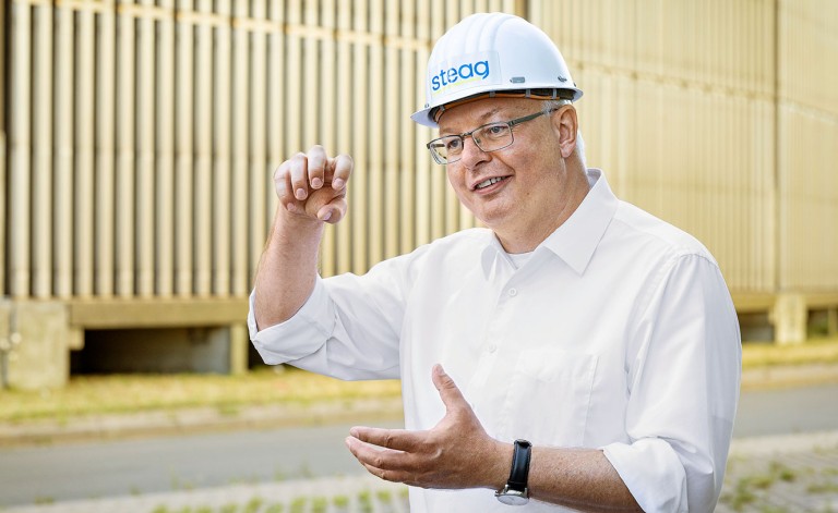 Ein Mann mit Brille und hellem Helm, auf dem „steag“ steht, steht vor einem großen Industriegebäude mit senkrechten Strukturen. Er trägt ein weißes Hemd und gestikuliert erklärend mit beiden Händen, während er lächelnd in eine Richtung blickt.