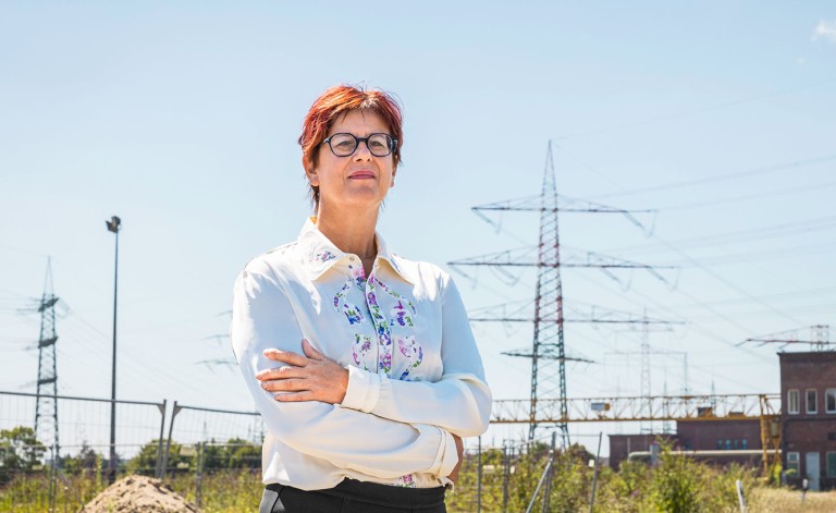 Eine Frau mit kurzen, rötlichen Haaren und Brille steht mit verschränkten Armen auf einer Baustelle oder einem Industriegelände. Sie trägt eine weiße Bluse mit buntem floralen Muster und blickt selbstbewusst in die Ferne. Im Hintergrund sind Strommasten, Bauzäune und ein Industriegebäude zu sehen. Der Himmel ist klar und blau.