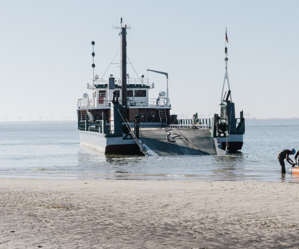 Das Bild zeigt ein großes Schiff, das an einem Ufer festgemacht ist. Das Schiff scheint ein Fährschiff oder ein Transportschiff zu sein, da es eine große, offene Ladefläche hat und eine Rampe heruntergelassen ist. Im Vordergrund sind mehrere Personen zu sehen, die eine orangefarbene Schlauchbootabdeckung oder eine ähnliche flexible Barriere ausrollen. Die Personen tragen Arbeitskleidung und scheinen eine Aufgabe im Zusammenhang mit dem Schiff oder der Barriere zu erfüllen. Im Hintergrund ist ein weites Gewässer zu sehen, wahrscheinlich ein Fluss oder eine Meeresbucht. Der Himmel ist klar und hell, was auf eine sonnige Tageszeit hinweist. Das Bild vermittelt einen Eindruck von Arbeit und Aktivität an einem Küstenort.