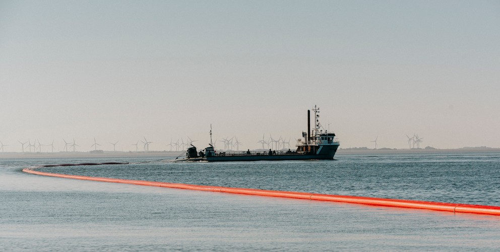 Das Bild zeigt ein Schiff, das in einem Gewässer vor Anker liegt. Hinter dem Schiff erstreckt sich eine lange, orangefarbene Rohrleitung über das Wasser bis zum Ufer. Im Hintergrund sind Windräder zu erkennen.