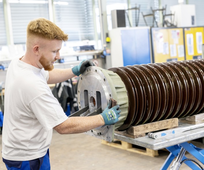 Das Bild zeigt einen Teil einer industriellen Produktionsstätte oder einer Werkstatt. Im Vordergrund ist ein großer, zylindrischer Gegenstand zu sehen, der auf einer Hebebühne steht. Dieser Gegenstand besteht aus einer Metallbasis, an der zahlreiche übereinander gestapelte, braune, isolierende Scheiben befestigt sind. Die Scheiben sind aus einem keramischen oder kunststoffartigen Material gefertigt und haben eine glatte, glänzende Oberfläche. Ein Arbeiter, der nur teilweise im Bild zu sehen ist, trägt Schutzhandschuhe und scheint den Gegenstand mit einem Werkzeug zu bearbeiten oder zu sichern. Im Hintergrund sind weitere Maschinen oder Werkzeuge erkennbar, die auf eine industrielle Umgebung hinweisen. Das Bild vermittelt einen Eindruck von Präzision und technischer Komplexität.