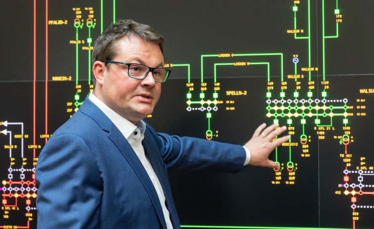 Ein Mann, Dr. Christoph Schneiders, steht in Geschäftskkleidung vor einem schwarzen Monitor mit bunt leuchtenden Schaltkreisen. Er deutet sprechend mit seiner linken Hand auf einen Bereich des Monitors.