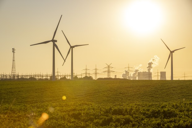 Windräder in der Abendsonne vor einer Umspannanlage, einem Kraftwerk und Strommasten im Hintergrund.