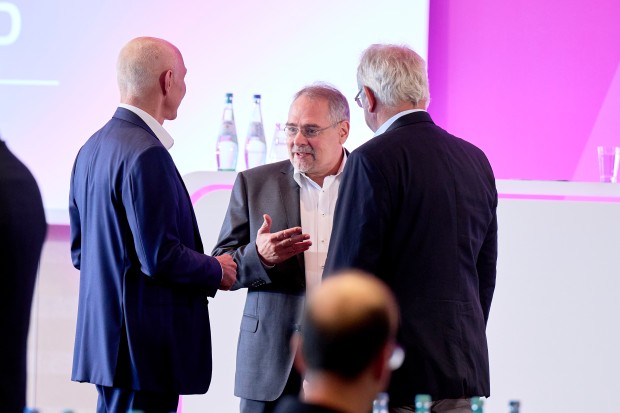 Drei Männer in Geschäftskleidung im Gespräch vor einem Podium.