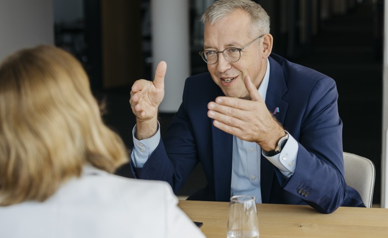 Gesprächssituation: Dr. Christoph Müller sitzt in Geschäftskleidung an einem Tisch. Im Vordergrund ist eine weitere Person von hinten erkennbar. Herr Dr. Müller gestikuliert in Richtung der Person.