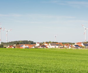 Ein grünes Feld im Vordergrund, im Hintergrund ein Dorf. Auf einigen Dächern befinden sich PV-Anlagen, dahinter Windräder. Die Sonne scheint, der Himmel ist wolkenfrei. 