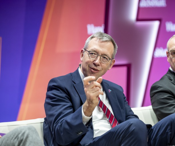 Das Bild zeigt Herrn Müller, den CEO von Amprion, in einem professionellen Umfeld. Er trägt eine Brille mit dünnem Metallrahmen, einen dunkelblauen Anzug, ein weißes Hemd und eine dezente Krawatte. Auf dem Revers seines Anzugs befindet sich das Amprion-Logo als kleines Abzeichen. Herr Müller lächelt leicht und gestikuliert mit der rechten Hand, als würde er etwas erklären. Am Ohr ist eine Mikrofonhalterung sichtbar, was darauf hinweist, dass er gerade spricht. Der Hintergrund ist in dynamischen Farben gehalten – Lila, Pink und Orange –, was auf eine moderne Veranstaltung oder ein Panel hinweist.