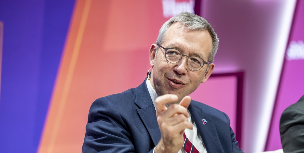 Das Bild zeigt Herrn Müller, den CEO von Amprion, in einem professionellen Umfeld. Er trägt eine Brille mit dünnem Metallrahmen, einen dunkelblauen Anzug, ein weißes Hemd und eine dezente Krawatte. Auf dem Revers seines Anzugs befindet sich das Amprion-Logo als kleines Abzeichen. Herr Müller lächelt leicht und gestikuliert mit der rechten Hand, als würde er etwas erklären. Am Ohr ist eine Mikrofonhalterung sichtbar, was darauf hinweist, dass er gerade spricht. Der Hintergrund ist in dynamischen Farben gehalten – Lila, Pink und Orange –, was auf eine moderne Veranstaltung oder ein Panel hinweist.