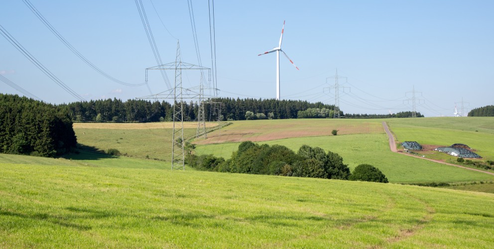 Das Bild zeigt eine Trasse in hügeliger Landschaft. Windräder sind am Horizont zu sehen.