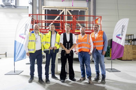 Fünf Männer stehen in einer Halle vor einer roten Scherenbühne. Zwei von ihnen tragen gelbe Sicherheitswesten, zwei orangefarbene und einer ist in traditioneller schwarzer Arbeitskleidung gekleidet. Im Hintergrund sind Werbefahnen und die Wände der Halle zu sehen.
