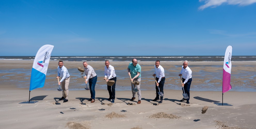 Sechs Männer in Hemden und Hosen stehen nebeneinander auf einem Sandstrand und halten Spaten in den Händen, als würden sie etwas ausgraben. Sie blicken alle in Richtung des Betrachters. Hinter ihnen erstreckt sich das Meer unter einem klaren blauen Himmel mit wenigen Wolken. Links und rechts im Bild sind jeweils eine Fahne mit dem Logo "amprion" zu sehen. Die Fahne auf der linken Seite ist blau und weiß, die auf der rechten Seite ist violett und weiß.