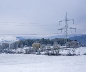 Das Bild zeigt eine winterliche Landschaft mit leicht verschneiten Feldern und sanften Hügeln. Mehrere Strommasten mit Hochspannungsleitungen durchziehen die Szenerie und bilden einen Kontrast zur natürlichen Umgebung. Im Vordergrund steht ein einzelner großer Strommast, dessen Leitungen sich weit über die verschneite Landschaft erstrecken.