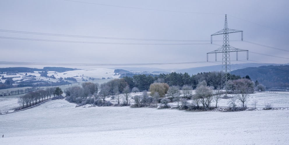 Das Bild zeigt eine winterliche Landschaft mit leicht verschneiten Feldern und sanften Hügeln. Mehrere Strommasten mit Hochspannungsleitungen durchziehen die Szenerie und bilden einen Kontrast zur natürlichen Umgebung. Im Vordergrund steht ein einzelner großer Strommast, dessen Leitungen sich weit über die verschneite Landschaft erstrecken.